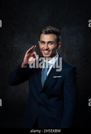 Ein junger Mann, der in einem Anzug lächelt, macht eine Handbewegung, alles ist in Ordnung. Vor einem dunklen Hintergrund. Stockfoto