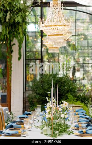 Bankett-Hochzeitstisch mit blauen Servietten, goldenem Besteck, Kristall, frischen Blumen und Kerzen. Hochzeitsdekorationen. Weicher, selektiver Fokus. Stockfoto