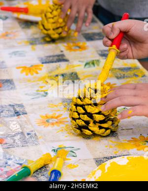 Nahaufnahme der Hände von Kindern, die in einer kreativen Werkstatt für Kunsthandwerk Kiefernzapfen mit gelber Farbe dekorieren Stockfoto