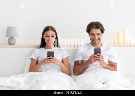 Lächelnde junge kaukasische Familie schaut sich Videos an, chattet in sozialen Netzwerken in Telefonen im Bett im Schlafzimmer Stockfoto