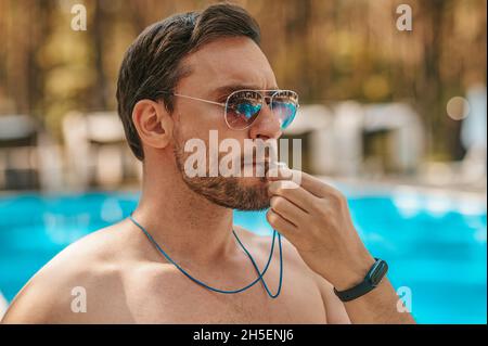 Männlicher Rettungsschwimmer mit Sonnenbrille in der Nähe des öffentlichen Schwimmbades Stockfoto