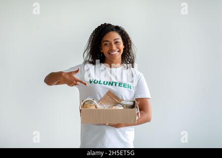 afroamerikanische Freiwillige hält und zeigt auf eine Kiste mit Lebensmittelspenden, posiert über hellem Studiohintergrund Stockfoto