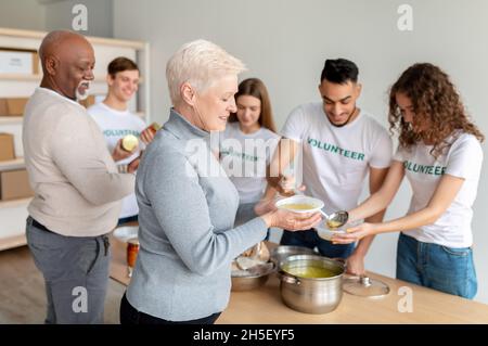 Glückliche ältere Frau erhielt warme Suppe im Büro der Wohltätigkeitsorganisation. Freiwilligengruppe hilft, ältere Menschen zu ernähren Stockfoto