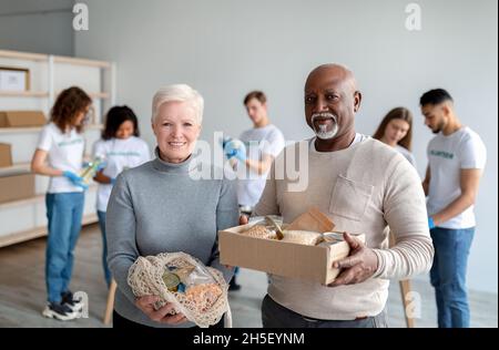 Glückliches gemischtes Seniorenpaar hält Spendennahrung in Kiste und Saitenbeutel und lächelt der Kamera im Charity Center zu Stockfoto