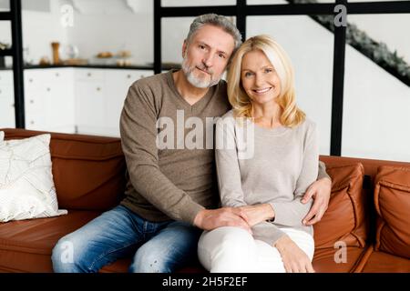 Fröhliches, älteres Paar, das lächelt und sich im gemütlichen Wohnzimmer umarmt Stockfoto
