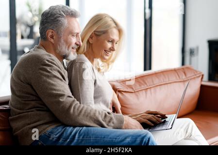 Ein verliebtes Paar mittleren Alters mit einem Laptop, der zu Hause auf der Couch sitzt. Fröhliches Seniorenpaar schaut sich gemeinsam Filme an, lächelt und umarmt sich im gemütlichen Wohnzimmer, surft im Internet und verbringt Zeit im Internet Stockfoto