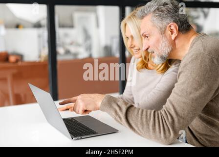 Fröhliches Paar mittleren Alters verbringt Freizeit online mit einem Laptop zu Hause. Hübscher grauhaariger Mann zeigt mit dem Finger auf den Bildschirm, schöne blonde Frau schaut zu und lacht. Seitenansicht Stockfoto