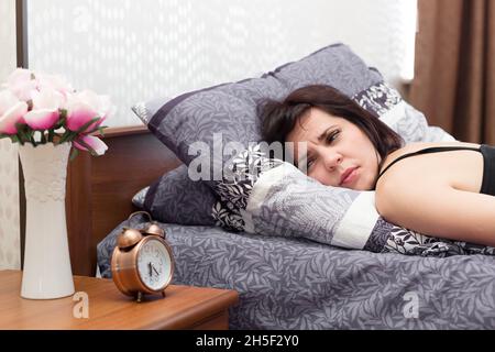 Die schlafende Frau öffnete ihre Augen mit Schwierigkeiten und blickt schmerzhaft auf den Wecker. Stockfoto