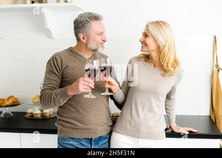 Ein romantisches Seniorenpaar verbringt den Abend zusammen, steht in der Küche mit Gläsern Rotwein, ein hübscher grauhaariger Mann und eine charmante Frau mittleren Alters schauen sich gegenseitig an und jubeln mit Gläsern Stockfoto