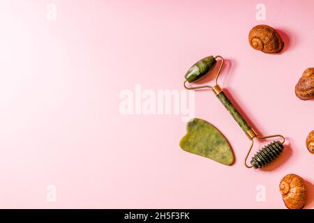 Ein grüner Jaderoller und ein Gesichtsmassage-Scrubber liegen auf einem weichen rosa Hintergrund. Stockfoto