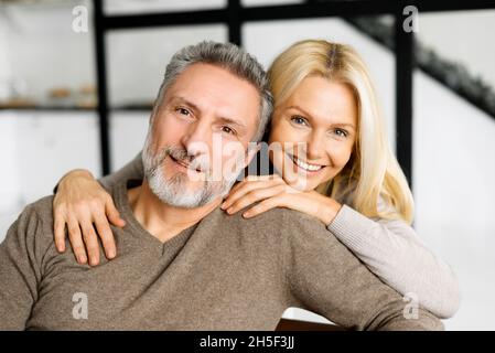 Liebe und Unterstützung Konzept. Porträt von Ehegatten mittleren Alters, glückliche, schöne Frau umarmt ihren grauhaarigen Mann, der auf der Couch sitzt, sie schauen auf die Kamera und lächeln Stockfoto