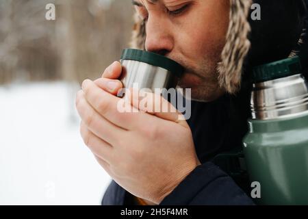 Ein Mann trinkt einen Kaffee aus einer Thermoskanne und wärmt seine Hände, steht auf dem Winter im Wald. Nahaufnahme Stockfoto