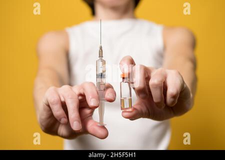 Die Hände halten eine Spritze mit Flüssigkeit und ein Fläschchen mit einem Impfstoff. Konzept der Impfung gegen Covid-19. Gelber Hintergrund. Nahaufnahme. Stockfoto