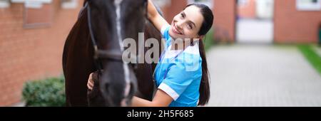 Lächelnde Tierärztin umarmt schwarzes Pferd in der Nähe von Ställen Stockfoto