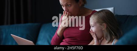 Schockiert überraschte Mutter und Tochter, die auf den Laptop-Bildschirm schauten, während sie auf der Couch saßen Stockfoto