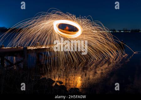 Leichte Malerei mit Stahlwolle auf einem Steg; pyrotechnische Darstellung bei Nacht mit Spiegelung im Wasser. Stockfoto