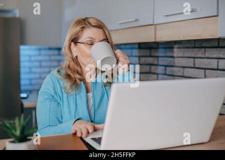 Eine Frau mittleren Alters trinkt Kaffee, während sie zu Hause mit ihrem Laptop Telearbeit führt. Porträt einer Geschäftsfrau. Arbeiten von zu Hause aus Stockfoto