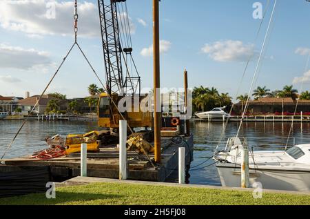 Reparatur von Ufermauer, Kran auf Lastkahn, sehr nah an angedocktem Segelboot, schwere Ausrüstung, Wartung, Florida, Punta Gorda, FL Stockfoto