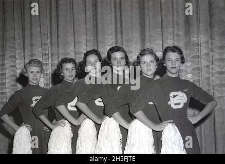 In den 1950er Jahren, historisch, gegen einen Vorhang, sechs amerikanische Cheerleadern in ihren Uniformen und halten ihre Bommel, die Hände auf Hüften stehend, in einer Schlange für ein Foto, USA. Cheerleader sind seit Jahrzehnten ein Teil des amerikanischen Mannschaftssports, sowohl Amateur- als auch Profisportler, insbesondere im Basketball und American Football, wo Mädchen spezielle Outfits tragen und ihre Bommel-Poms winken, während sie ihre Teams anfeuern und unterstützen. In dieser Zeit wurde der erste Brief der High School, an der die Cheerleader teilnahmen, oft auf die Outfits gestickt - wie wir hier den Buchstaben G sehen - in der Mitte einer Megaphon-Form. Stockfoto