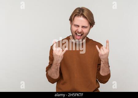 Porträt eines Mannes mit Bart, der Sweatshirt trägt, mit Rock and Roll-Zeichen-Handbewegung, schreiend, mit aufgeregt Gesichtsausdruck. Innenaufnahme des Studios isoliert auf grauem Hintergrund. Stockfoto