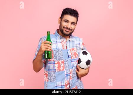 Porträt eines jungen erwachsenen bärtigen Mannes, der mit einer Flasche Alkohol und einem Fußballball in den Händen steht, die Kamera anschaut, das Spiel mit Bier beobachtet. Innenaufnahme des Studios isoliert auf rosa Hintergrund. Stockfoto