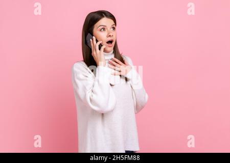 Seitenansicht eines überraschten weiblichen Sprechtelefons, das Nachrichten hörte, den Mund offen hielt und einen weißen Pullover im lässigen Stil trug. Innenaufnahme des Studios isoliert auf rosa Hintergrund. Stockfoto