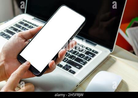 Man Hand drückt das Smartphone mit einem leeren Bildschirm für die Gestaltung oder Werbung auf dem Schreibtisch und den Laptop-Computer auf dem Arbeitstisch. Stockfoto