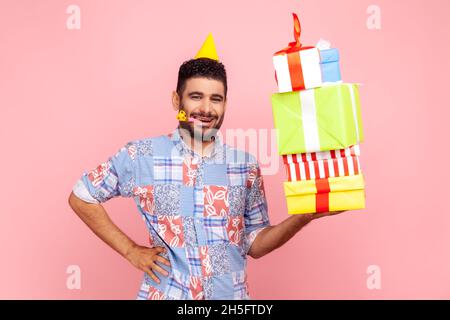 Glücklicher, positiver Mann mit Bart, der Geschenkboxen trägt, Party-Horn bläst, Hand an Hüfte hält und mit zufriedenem Ausdruck die Kamera anschaut. Innenaufnahme des Studios isoliert auf rosa Hintergrund. Stockfoto