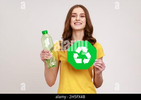 Glücklich zufrieden Teenager Mädchen mit braunen Haaren in gelben T-Shirt in der Hand halten Recycling Schild und Plastikflasche, Mülltrennung. Innenaufnahme des Studios isoliert auf grauem Hintergrund. Stockfoto