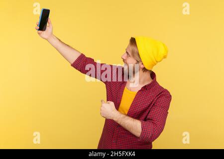 Porträt eines bärtigen Mannes mit Hipster in Beanie-Hut und kariertem Hemd, der während eines Videoanrufs spricht, Online-Gespräche auf dem Mobiltelefon führt, Selfie macht. Innenaufnahme des Studios isoliert auf gelbem Hintergrund. Stockfoto