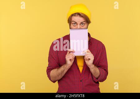 Porträt eines bärtigen Mannes mit Hipster in Beanie-Hut und kariertem Hemd, das die Hälfte des Gesichts mit einem Notizbuch aus Papier bedeckt und die Kamera anschaut. Innenaufnahme des Studios isoliert auf gelbem Hintergrund. Stockfoto