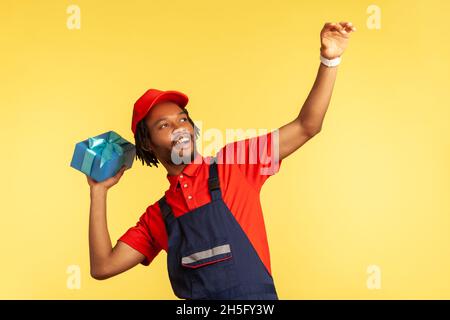 Porträt eines lächelnden glücklichen männlichen Kuriers in professioneller blauer Uniform und roter Mütze, der Geschenke wirft, schnelle Lieferung. Innenaufnahme des Studios isoliert auf gelbem Hintergrund. Stockfoto