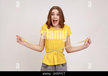 Portrait glücklich schlanke Frau in gelben Casual-Stil T-Shirt hält auf Taille Maßband und Blick auf die Kamera mit Zufriedenheit und Aufregung. Innenaufnahme des Studios isoliert auf grauem Hintergrund. Stockfoto