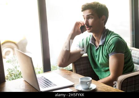 Seitenansicht Porträt eines Angestellten mit grünem T-Shirt, einem Telefongespräch mit schockiert erstauntem Gesichtsausdruck, das online am Laptop arbeitet. Innenaufnahme in der Nähe des großen Fensters, Café-Hintergrund. Stockfoto