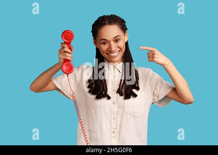 Porträt einer lächelnden zufriedenen Frau mit schwarzen Dreadlocks halten zeigt auf rote retro Telefon, Call Center. Tragen weißes Hemd. Innenaufnahme des Studios isoliert auf blauem Hintergrund. Stockfoto
