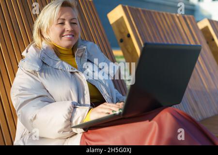 Eine wunderschöne kaukasische Frau mittleren Alters, die an einem Laptop auf der Holzbank auf der Straße sitzt. Stockfoto