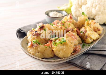 Geröstete Blumenkohlröschen mit Ei und Schinken auf einem grauen Teller und Quark-Dip auf einer Serviette und einem Holztisch, gesundes Gemüsegericht für eine kohlenhydratarme Ernährung, Stockfoto