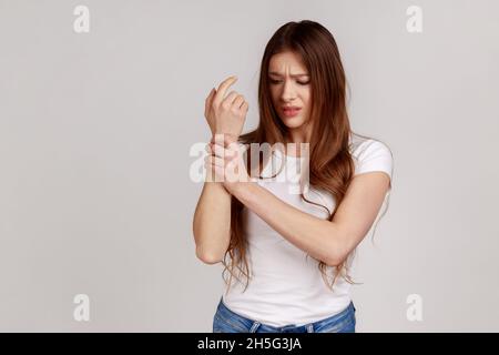 Unzufrieden ungesunde Frau, die mit Schmerzen steht, schmerzende Handgelenke massiert, Handverletzungen oder Verstauchungen leidet und weißes T-Shirt trägt. Innenaufnahme des Studios isoliert auf grauem Hintergrund. Stockfoto