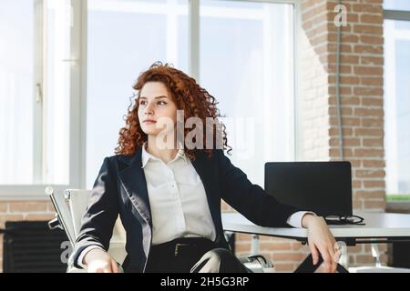 Porträt einer schönen erfolgreichen jungen Geschäftsfrau mit Rot Lockiges Haar im Büroinneren sitzt an einem Tisch In der Nähe des Fensters Stockfoto