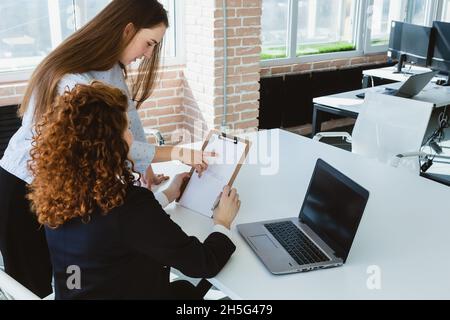 Schöne junge Brünette Frau und rothaarige Frau reden und diskutieren Arbeitsmomente mit Laptop im Büro. Stockfoto