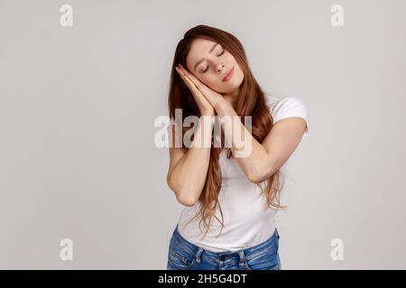 Porträt einer schönen Frau, die auf ihren Handflächen schläft, ein bequemes Nickerchen macht und sich ausruht, sich niederlässt und weißes T-Shirt trägt. Innenaufnahme des Studios isoliert auf grauem Hintergrund. Stockfoto