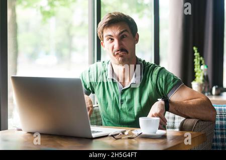 Porträt eines Mitarbeiters, der ein grünes T-Shirt trägt, die Augen kreuzt, herumtollt und Spaß hat, am Arbeitsplatz vor dem Laptop sitzt. Innenaufnahme in der Nähe des großen Fensters, Café-Hintergrund. Stockfoto