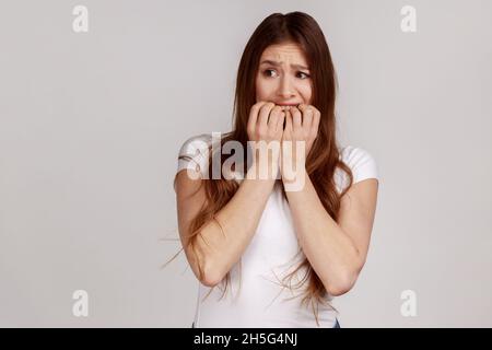 Porträt einer nervösen Frau mit langen dunklen Haaren, beißende Nägel und schauend erschrocken, erschrocken über Probleme, Angststörungen, das Tragen eines weißen T-Shirts. Innenaufnahme des Studios isoliert auf grauem Hintergrund. Stockfoto