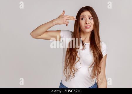 Porträt einer verzweifelten Frau, die mit dem Finger auf den Kopf zeigt, sich selbst erschießt, Selbstmordgesten macht, um Depressionen zu stoppen, in einem weißen T-Shirt. Innenaufnahme des Studios isoliert auf grauem Hintergrund. Stockfoto