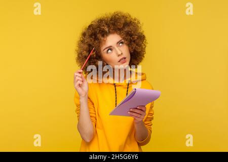 Schöne nachdenkliche Frau mit Afro-Frisur, die Notizblock und Bleistift in den Händen hält und über Pläne für das Startup nachdenkt, trägt einen lässigen Hoodie. Innenaufnahme des Studios isoliert auf gelbem Hintergrund. Stockfoto