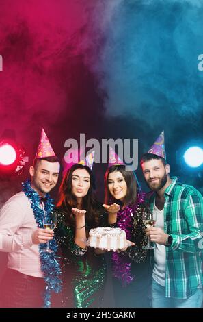 Geburtstagsfeier. Eine Gruppe junger Menschen in Geburtstagskappen mit einem Kuchen und Gläsern Wein oder Champagner in den Händen schauen auf die Kamera auf einer da Stockfoto