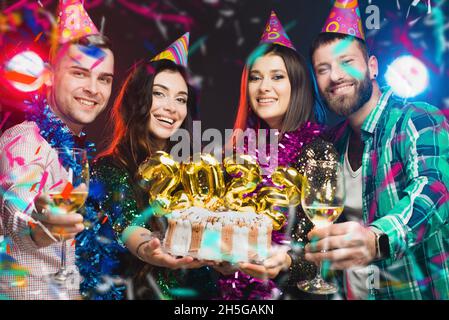 Eine Gruppe junger Menschen in Geburtstagsmützen mit einem Kuchen in den Händen mit den Zahlen 2022 schauen vor dunklem Hintergrund mit Lig auf die Kamera Stockfoto
