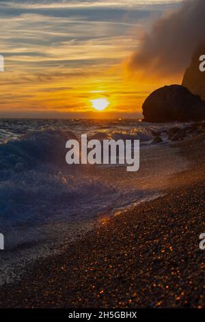 Sonnenuntergang und Sturm auf dem Meer. Große Wellen gegen die untergehende Sonne. Ein Sommersturm im Meer. Schönes Meeresspray mit Schaumbrüchen an der felsigen Küste. Vert Stockfoto