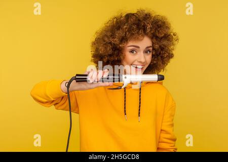 Frau hält Lockenstab, macht perfekte Afro-Frisur, Bügeln von Locken, Pflege mit professionellem Werkzeug, trägt lässigen Stil Hoodie. Innenaufnahme des Studios isoliert auf gelbem Hintergrund. Stockfoto