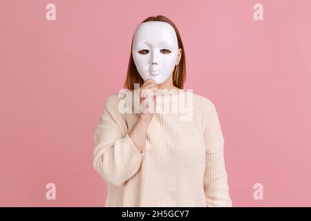 Porträt einer unbekannten anonymen Frau, die ihr Gesicht mit einer weißen Maske bedeckt, ihre wahre Persönlichkeit, Anonymität, versteckt und weißen Pullover trägt. Innenaufnahme des Studios isoliert auf rosa Hintergrund. Stockfoto